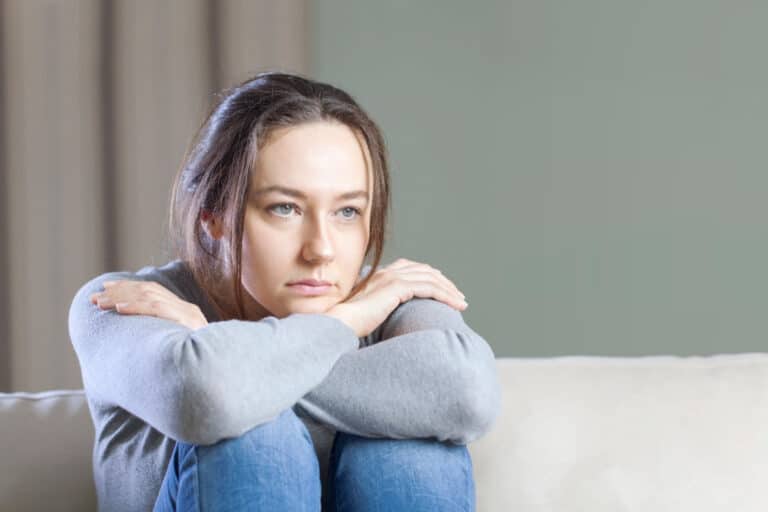 Woman in grey shirt sits with her knees pulled to her chest looking forward in thought.