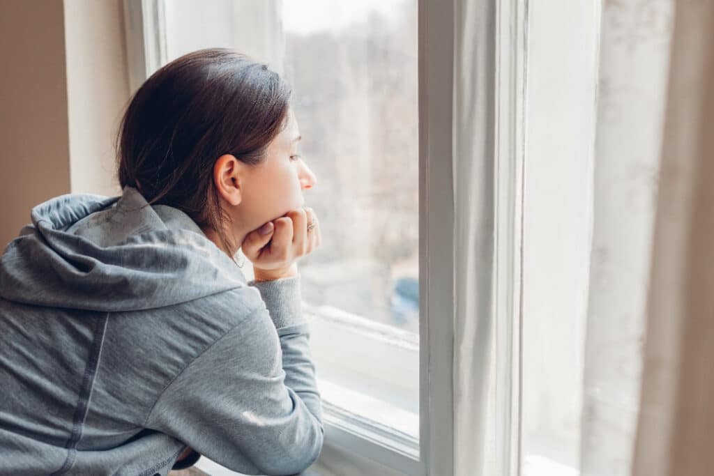 Woman in grey hoodie sweatshirt looking out a window, thinking.
