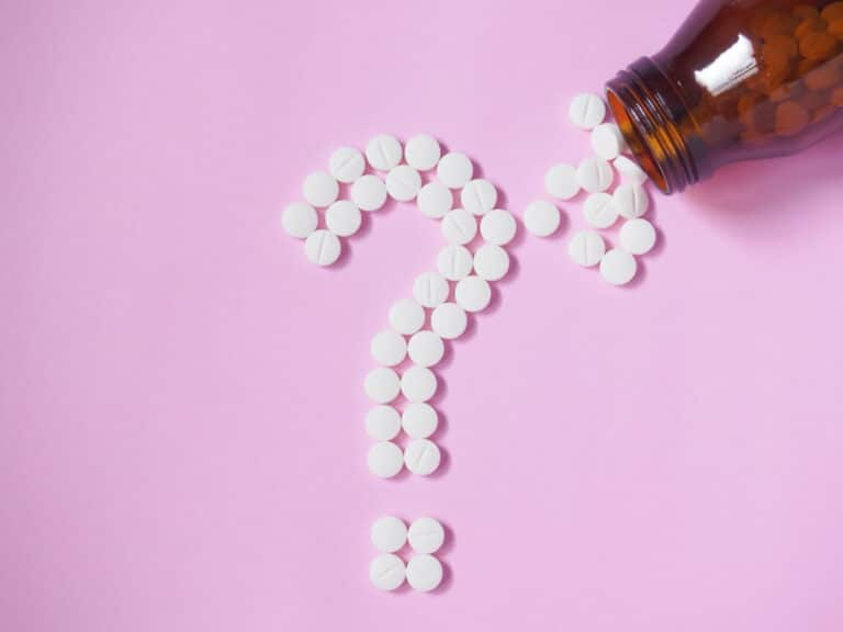 White, round pills coming out of a bottle and forming a question mark on a pink background.