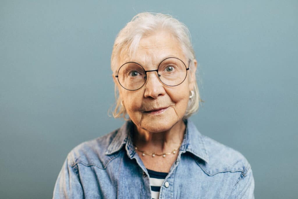 An older woman with white hair and round glasses. She's dressed stylishly in a denim shirt, and she's looking straight at the camera.