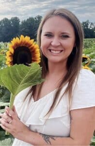 Victoria is smiling at the camera as she hold a sunflower. 